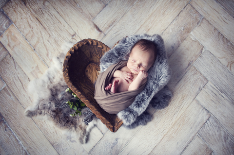 Newborn baby boy wrapped in brown wrap on gray furry rabbit blanket in wooden bowl in the shape of heart on wooden floor